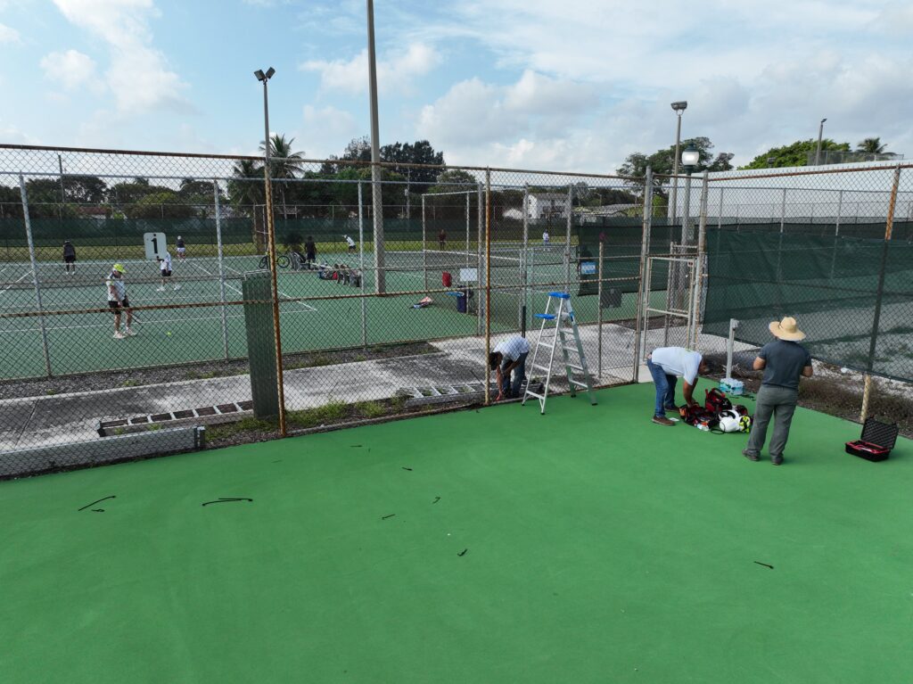 A tennis court with people and equipment beside it under a cloudy sky.