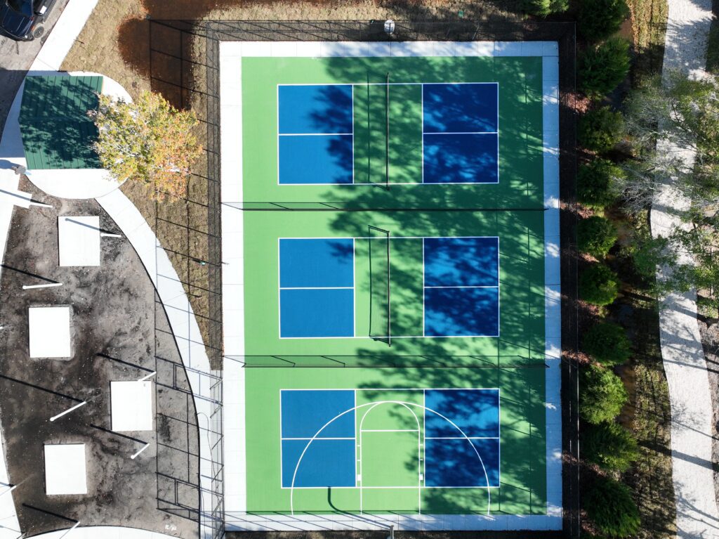 Aerial view of a tennis court with six blue playing areas and green surroundings.