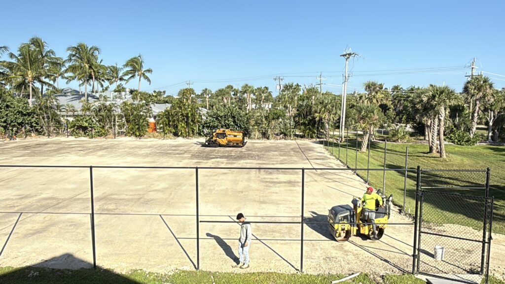 A fenced sports court with workers and a yellow vehicle on a sunny day.