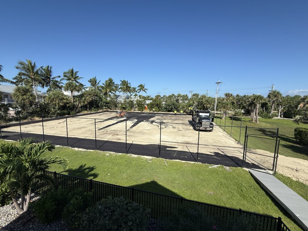 A fenced sandy volleyball court with palm trees and a clear blue sky.