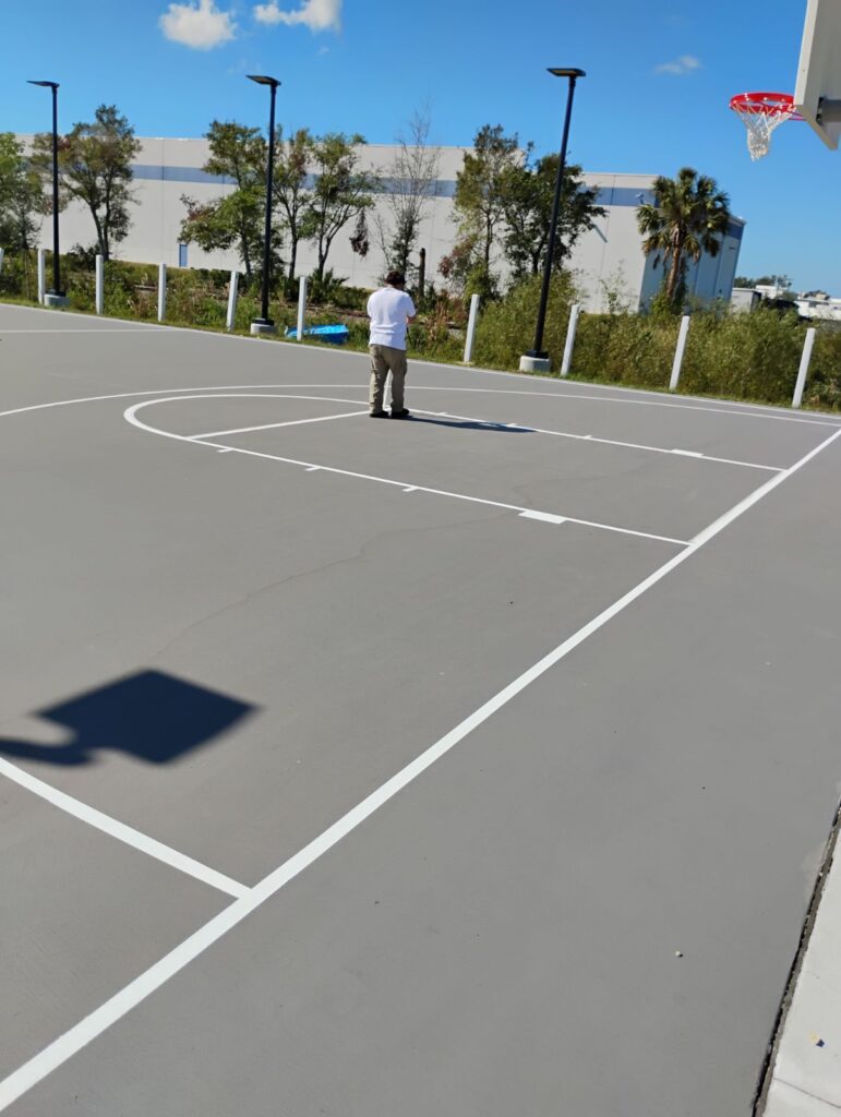 Person standing on an empty outdoor basketball court on a sunny day.